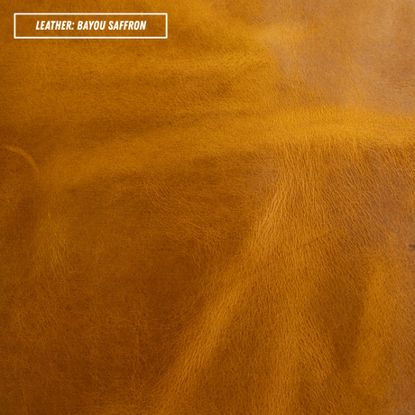 A close-up texture shot of smooth leather in the color 'Bayou Saffron', showing a warm golden-brown hue with subtle natural grain and folds. A white label in the top-left corner reads 'LEATHER: BAYOU SAFFRON'.
