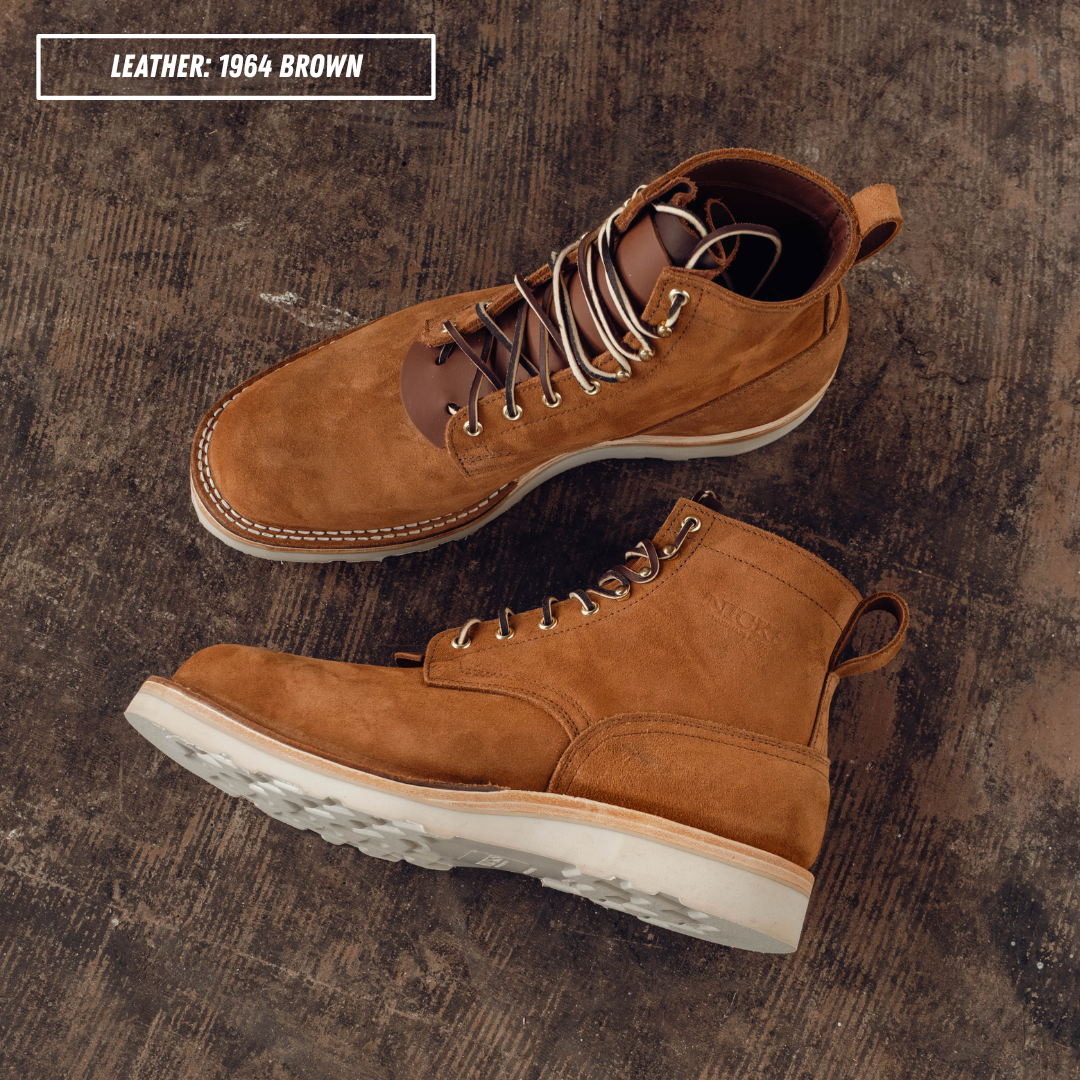 A pair of  brown leather lace-up boots arranged on a dark textured concrete surface, with a text label reading 'LEATHER: 1964 BROWN' in the top left corner.