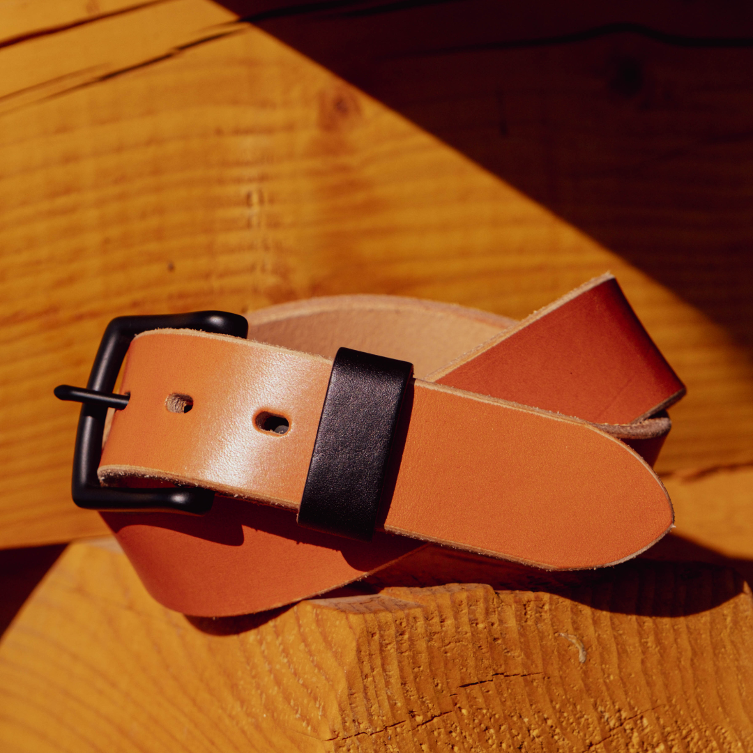 A tan leather belt with a black buckle and black keeper loop, coiled on a rustic wooden surface.