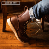 Model wearing brown Moctoepro while sitting in chair. Label on top left corner that reads "LEATHER: 1964 BROWN."