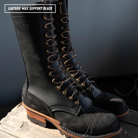 A pair of black leather lace-up boots with tan laces and brown stacked soles, shown on a wooden crate with grey background and a text label in the top left corner.