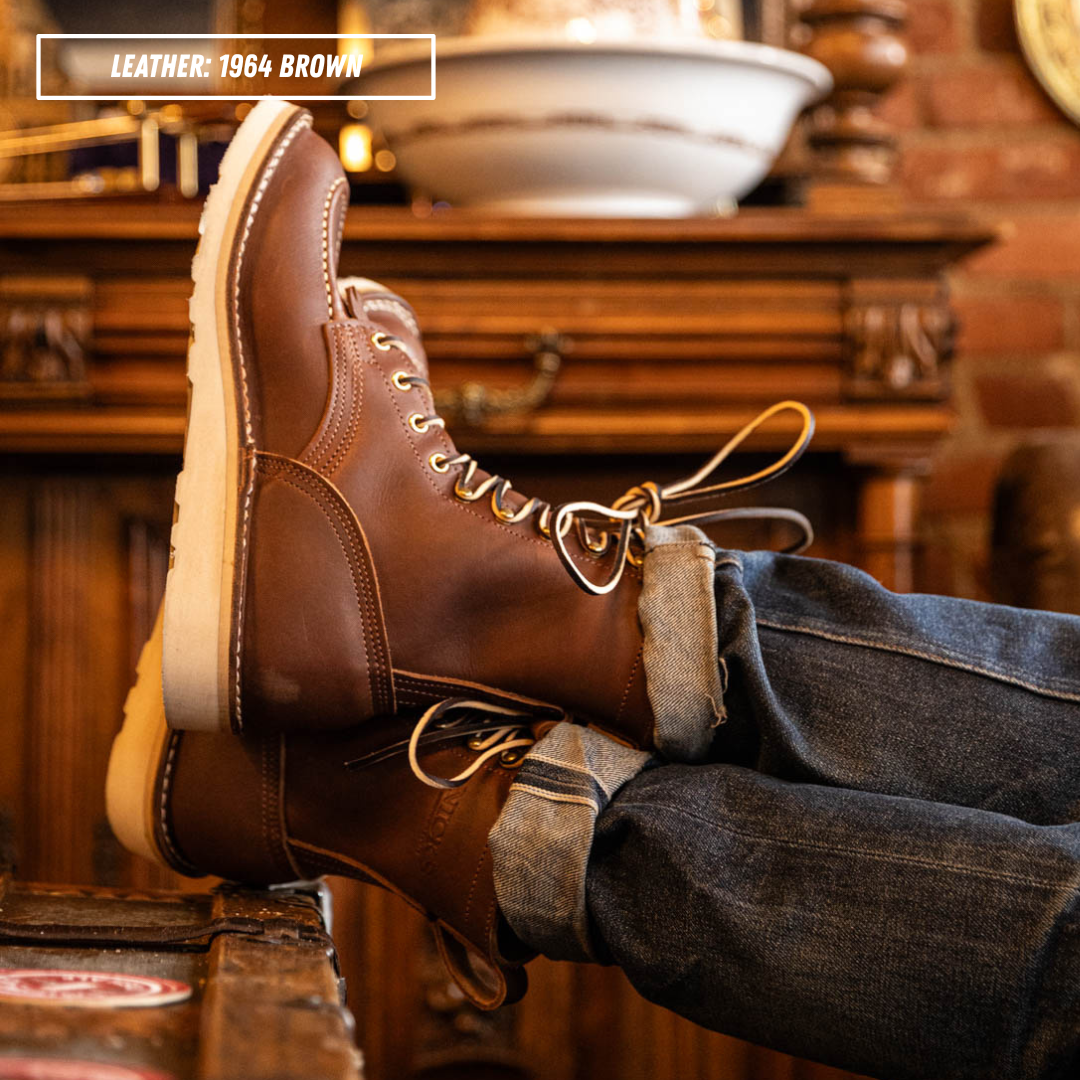 Model wearing brown Moctoepro while sitting in chair kicking feet up on table. Label on top left corner that reads "LEATHER: 1964 BROWN."
