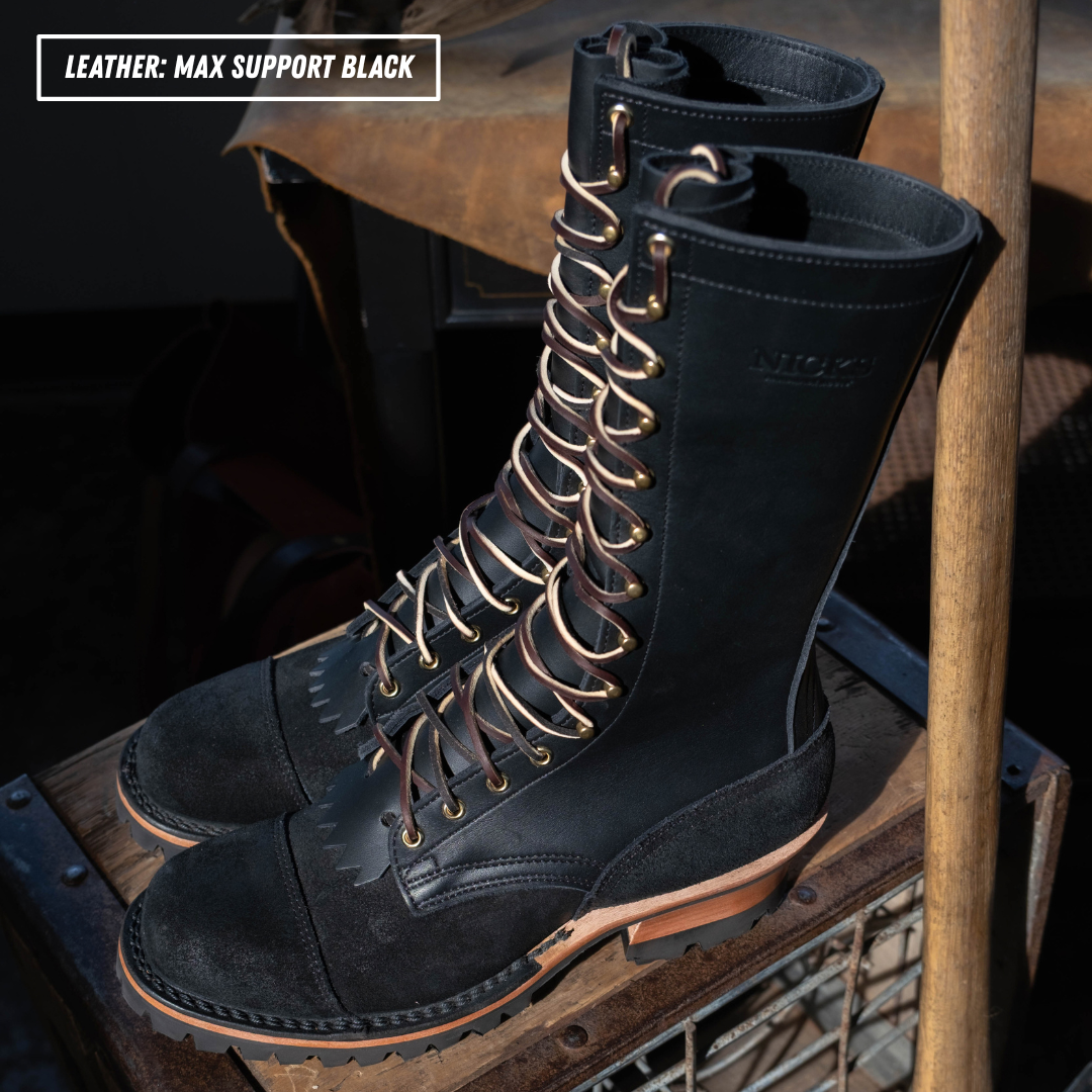 A pair of tall black leather lace-up boots displayed on a rustic wooden crate in a workshop setting. The boots feature decorative laces with gold-toned metal loops and a tan outsole.