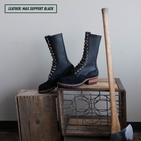 A pair of tall black leather lace-up boots displayed on rustic wooden crates, with a wooden-handled tool leaning against the crate. A text label in the top-left corner reads 'LEATHER: MAX SUPPORT BLACK'.
