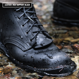 A close-up of a black leather lace-up boot with water droplets on its toe and sides, standing in shallow wet leaves and rocks. A second black boot is blurred in the background.