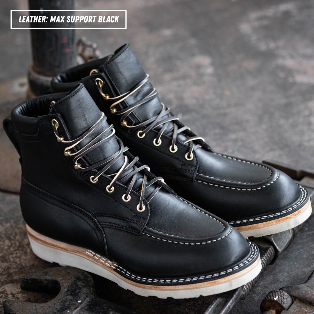 A pair of black leather lace-up ankle boots with white stitching and white soles, resting on a dark textured metal surface. A text overlay in the top-left corner reads 'LEATHER: MAX SUPPORT BLACK'.