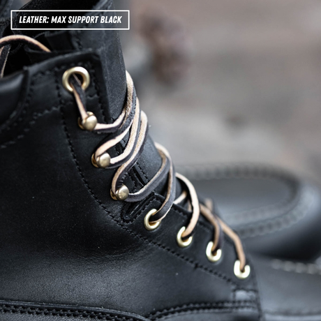 Close-up of a black leather lace-up boot with metallic eyelets and tan laces, with a text label in the top-left corner reading 'LEATHER: MAX SUPPORT BLACK'.