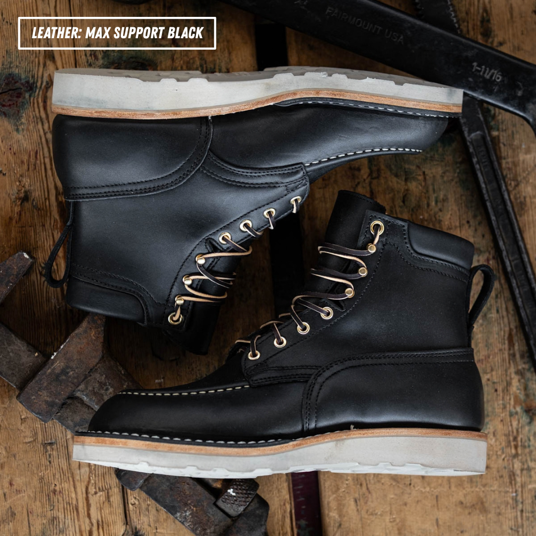 A pair of black leather lace-up work boots arranged on a rustic wooden surface with tools around them. Text in the top-left corner reads 'LEATHER: MAX SUPPORT BLACK'.