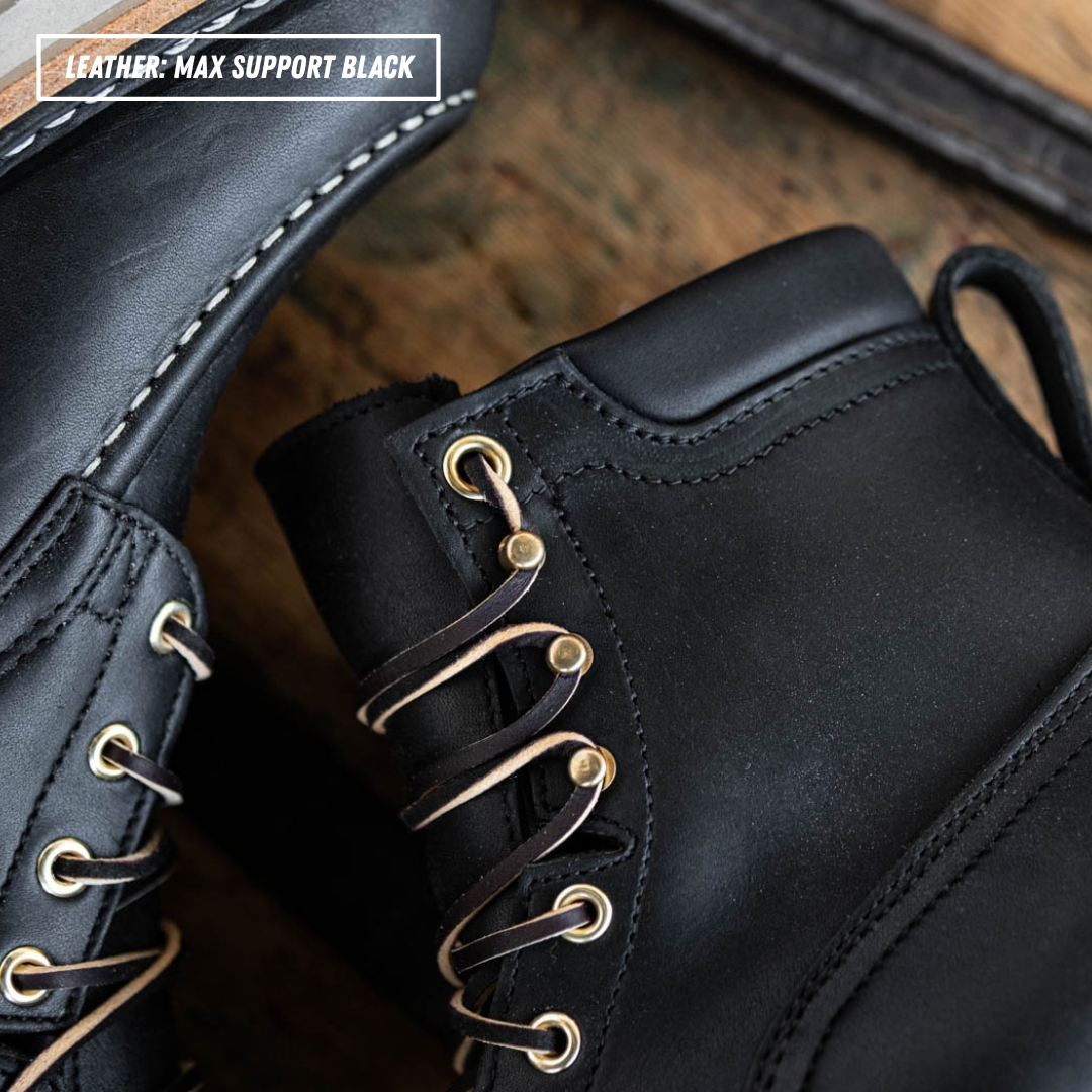 Close-up of a black leather lace-up boot with gold-tone eyelets and laces, shown on a rustic wooden surface. A second boot is partially visible at left, and a text overlay in the top left reads 'LEATHER: MAX SUPPORT BLACK'.