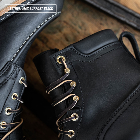 Close-up of a black leather lace-up boot with gold-tone eyelets and laces, shown on a rustic wooden surface. A second boot is partially visible at left, and a text overlay in the top left reads 'LEATHER: MAX SUPPORT BLACK'.