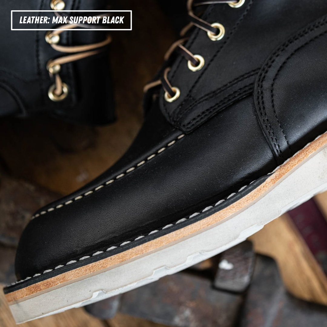 Close-up of a black leather lace-up boot with white contrast stitching, gold eyelets, and a white midsole over a brown outsole. A second boot is partially visible in the background.