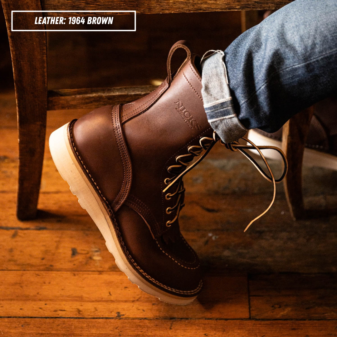 A brown leather lace-up work boot worn on a foot with cuffed blue jeans, photographed in a rustic indoor setting with wooden furniture and floorboards. A text label in the top left reads 'LEATHER: 1964 BROWN'.