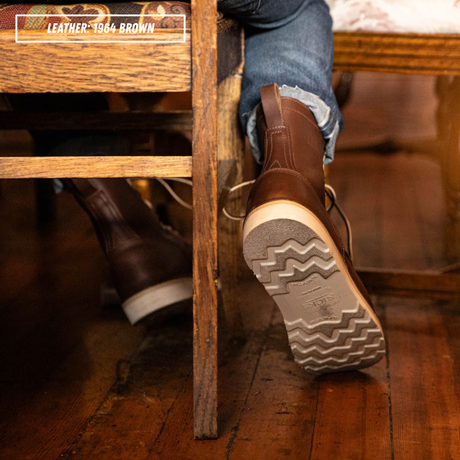A person wearing brown leather ankle boots with blue jeans, standing on a wooden floor under a wooden bench. One boot is shown from the side and the other from the sole, with a text overlay in the top left that reads 'LEATHER - 1964 BROWN'.