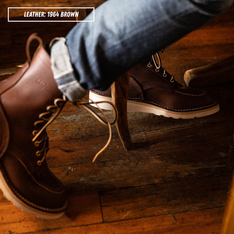 A close-up lifestyle shot of a person wearing a pair of brown leather lace-up boots with blue jeans, standing on a rustic wooden floor. A text overlay in the top-left corner reads 'LEATHER: 1964 BROWN'.