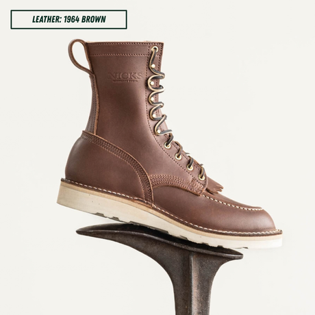 A single brown leather lace-up boot shown on a dark display stand against a light gray background, with a text label in the top-left corner reading 'LEATHER: 1964 BROWN'. The boot has tan stitching, metal eyelets, a pull loop, and a thick off-white sole.