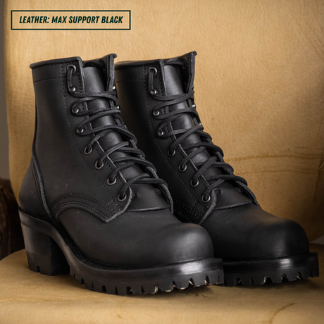 A pair of black leather lace-up boots with chunky lug soles and low block heels, displayed on a tan textured surface against a matching tan background. A text label in the top-left corner reads 'LEATHER: MAX SUPPORT BLACK'.