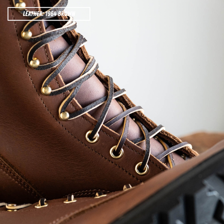 Close-up of a brown leather lace-up boot with dark brown laces threaded through brass eyelets, with a text label reading 'LEATHER: 1964 BROWN' in the top-left corner.