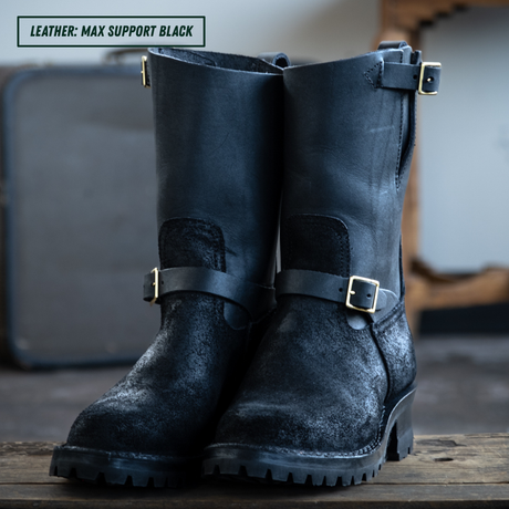 A pair of tall black leather boots with distressed textures and brass buckle straps, displayed on a wooden surface with a blurred background.