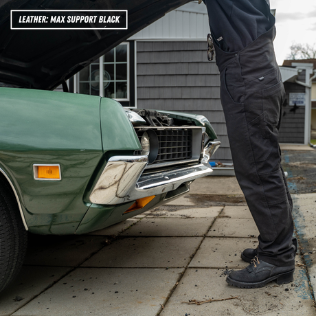 A person wearing black leather work pants stands beside an open green car hood in a driveway. The text overlay at the top left reads 'LEATHER: MAX SUPPORT BLACK'.