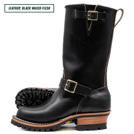 A single black leather mid-calf boot with gold-tone buckle details and a brown stacked heel, shown on a white background with a second boot showing the sole behind it. A text label in the top left reads 'LEATHER: BLACK WAXED FLESH'.