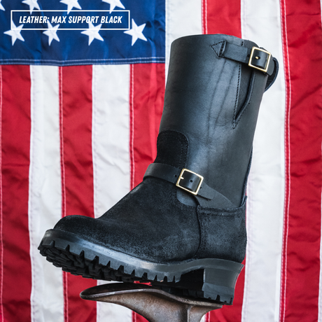A black leather mid-calf boot with buckle straps, shown standing on a metal stand in front of an American flag background. A text overlay in the top left reads 'LEATHER; MAX SUPPORT BLACK'.