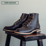 A pair of dark brown leather lace-up boots with tan laces and gold-toned eyelets, displayed on a rustic wooden stool against a light gray wall. A text label in the top left reads 'LEATHER: GALLUN BLACK'.