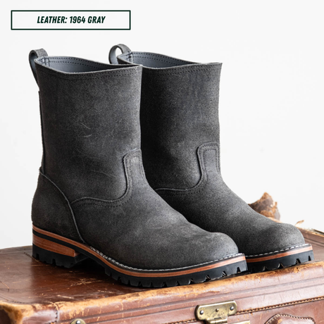 A pair of dark gray suede mid-calf boots with brown lugs, displayed on a vintage brown leather suitcase against a white background. A text overlay in the top left reads 'LEATHER: 1964 GRAY'.