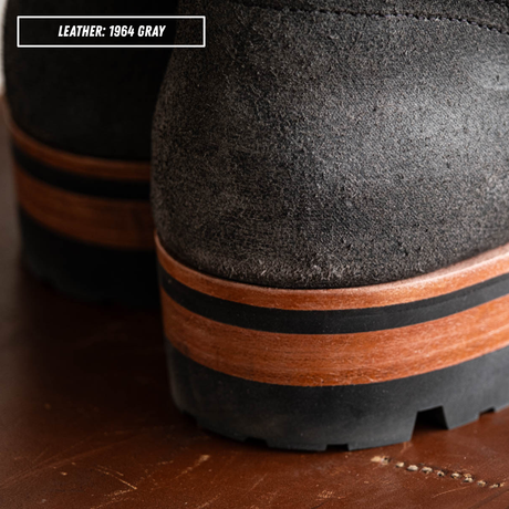 Close-up of the heel of a black leather boot in 1964 gray, showing a wood-look brown midsole band and a black rubber outsole on a brown wooden surface.
