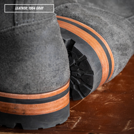 Close-up of a gray suede boot with a chunky black lug sole featuring a distinctive brown wood-look stripe, resting on a rustic wooden surface. A text overlay in the top left reads 'LEATHER: 1964 GRAY'.
