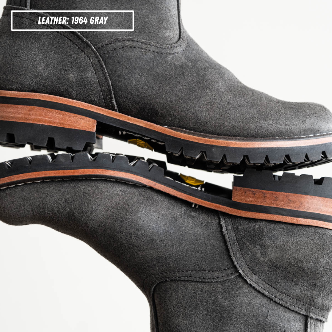 A close-up studio shot of a pair of dark gray leather boots stacked together to show the sole and side profile. A text label in the top left reads 'LEATHER: 1964 GRAY'.