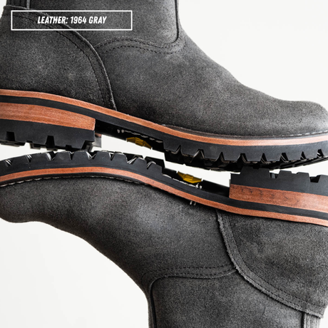 A close-up studio shot of a pair of dark gray leather boots stacked together to show the sole and side profile. A text label in the top left reads 'LEATHER: 1964 GRAY'.