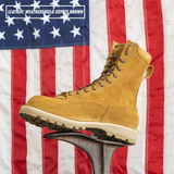 A tan suede lace-up work boot displayed on a metal stand in front of a blurred American flag. A white label in the top-left reads 'LEATHER: WEATHERSHIELD COYOTE BROWN'.