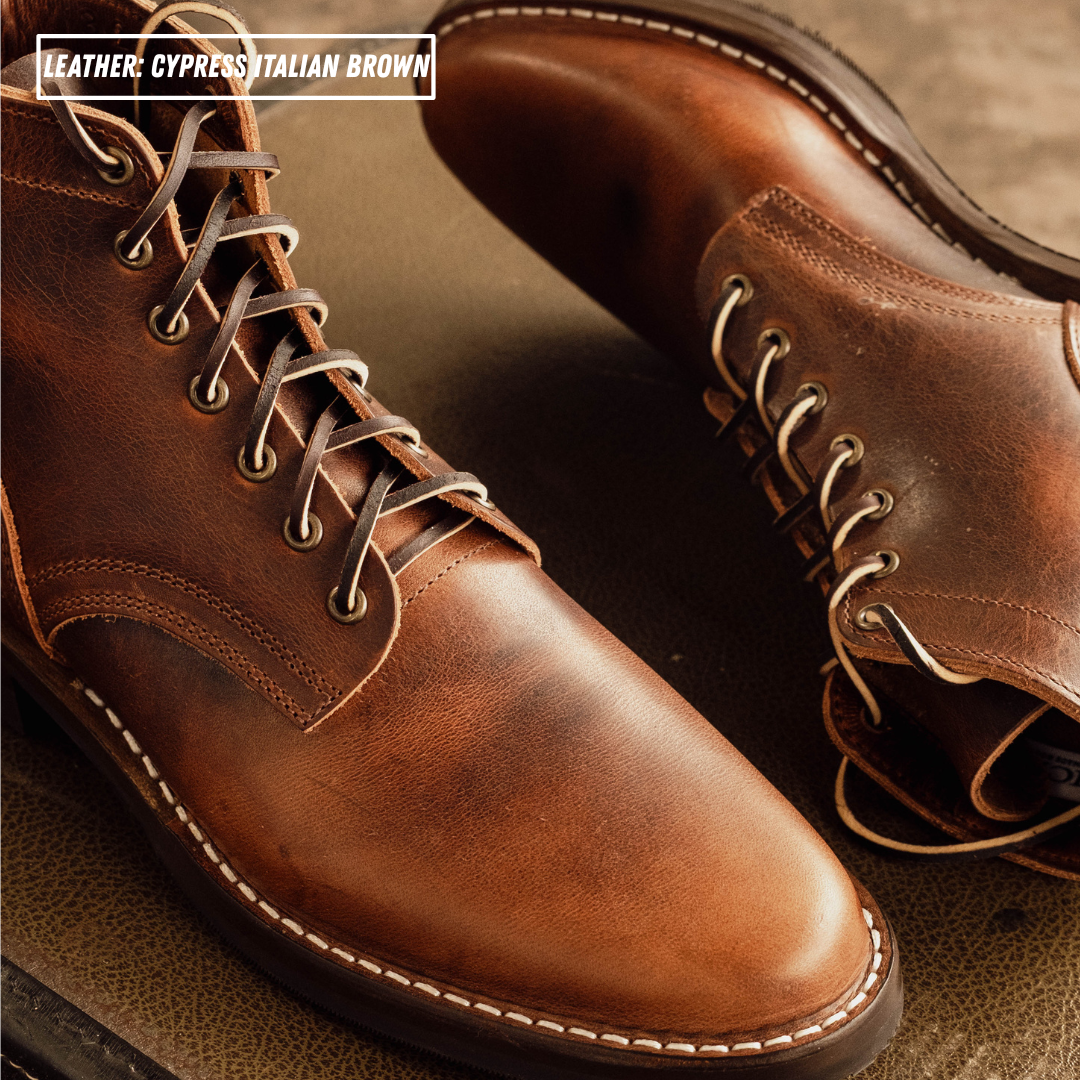A close-up of a pair of leather lace-up boots in a warm brown shade, with white contrast stitching and metal eyelets. Text in the top-left corner reads 'LEATHER: CYPRESS ITALIAN BROWN'.