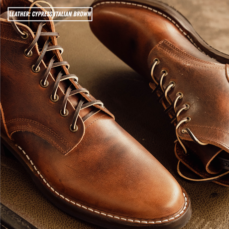A close-up of a pair of leather lace-up boots in a warm brown shade, with white contrast stitching and metal eyelets. Text in the top-left corner reads 'LEATHER: CYPRESS ITALIAN BROWN'.