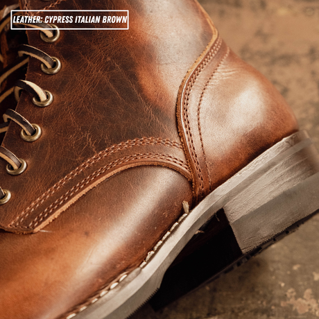 Close-up of a brown leather lace-up boot showing the side panel, stitching, eyelets, and part of the sole, with text reading 'LEATHER - CYPRESS ITALIAN BROWN'.