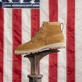 A single coyote brown suede work boot with a pull tab and lace-up front, displayed on a metal anvil in front of a red, white, and blue American flag. A text overlay in the top left reads 'LEATHER WEATHERSHIELD COYOTE BROWN'.