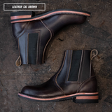 A pair of dark brown leather Chelsea boots with black elastic side panels and a tan welt, arranged on a dark rustic wooden surface. A text overlay in the top left reads 'LEATHER: CXL BROWN'.