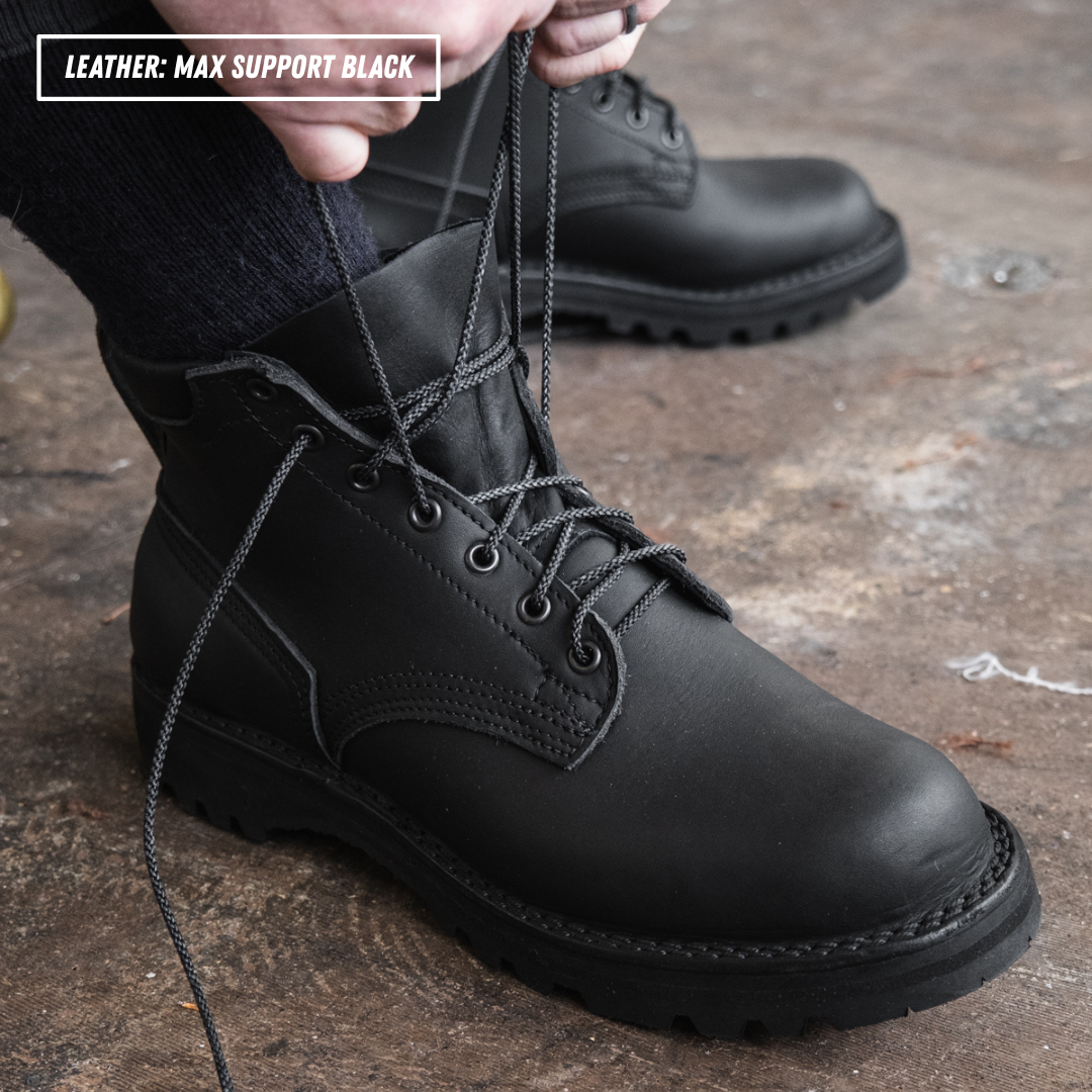 A close-up of a person wearing and tying a black leather lace-up boot, with a second matching boot blurred in the background on a rustic concrete floor. A text overlay in the top left reads 'LEATHER: MAX SUPPORT BLACK'.