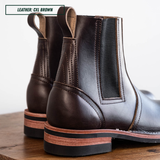 A pair of dark brown leather Chelsea boots with elastic side panels, shown from a close-up rear three-quarter angle on a wooden surface against a white background. A text overlay in the top-left corner reads 'LEATHER: CXL BROWN'.