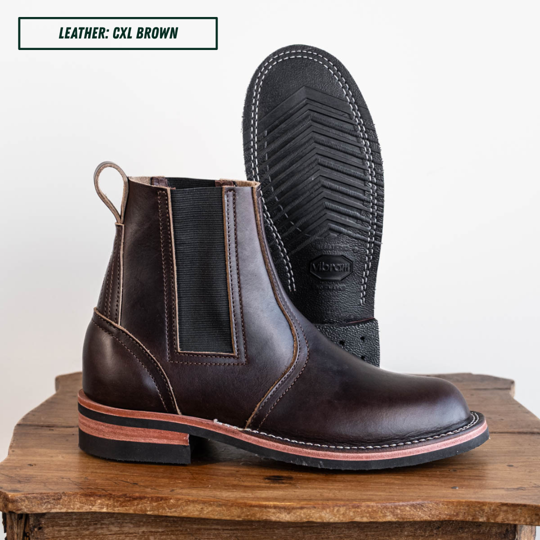 A pair of dark brown leather Chelsea boots with black elastic side panels, shown on a rustic wooden block against a white background. One boot is standing upright to show the side profile, and the other is turned upside down to display the black treaded sole.