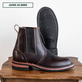 A pair of dark brown leather Chelsea boots with black elastic side panels, shown on a rustic wooden block against a white background. One boot is standing upright to show the side profile, and the other is turned upside down to display the black treaded sole.