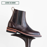 A dark brown leather Chelsea boot with a black elastic side panel, displayed on a black metal riser against a white background. A text label in the top-left corner reads 'LEATHER: CXL BROWN'.
