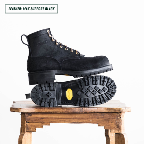 All black leather boots with lug soles. Boots sitting on wooden table, one laying down showing outsole and the other sitting side profile on top. Top left reads "LEATHER: MAX SUPPORT BLACK."