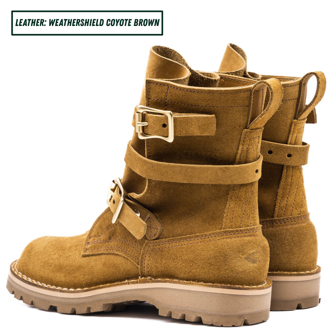 A pair of mid-calf boots in a warm coyote brown suede-like leather, featuring double buckle straps on the sides and a rugged lug sole. Boots sitting together facing back left.
