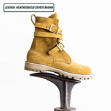 A pair of mid-calf boots in a warm coyote brown suede-like leather, featuring double buckle straps on the sides and a rugged lug sole. One boot is sitting on boot jack.