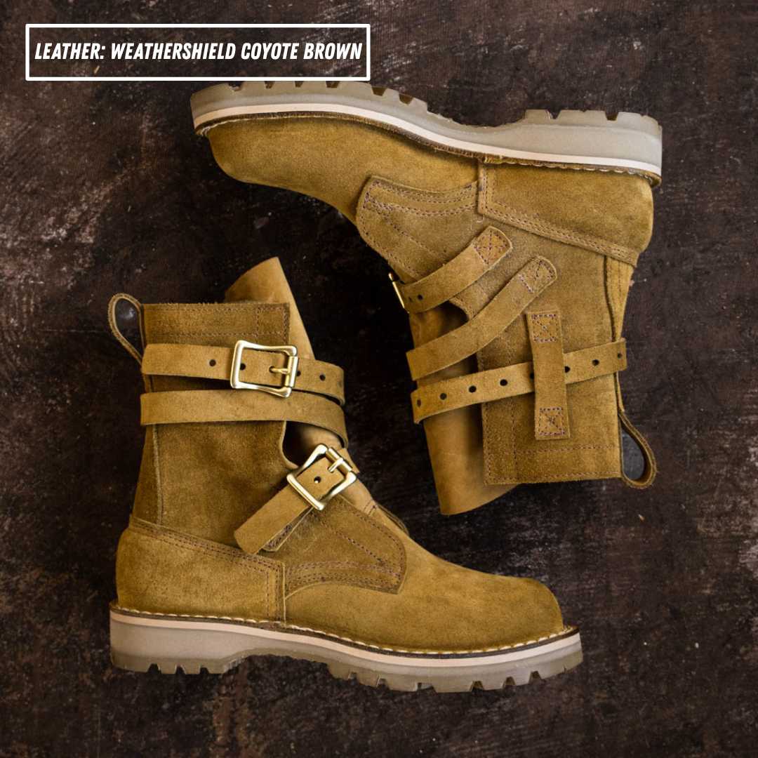 A pair of ankle-high boots in coyote brown suede, displayed on a dark textured background. One boot is shown from the side, and the other is angled to show the side profile and buckle straps.
