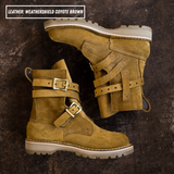 A pair of ankle-high boots in coyote brown suede, displayed on a dark textured background. One boot is shown from the side, and the other is angled to show the side profile and buckle straps.