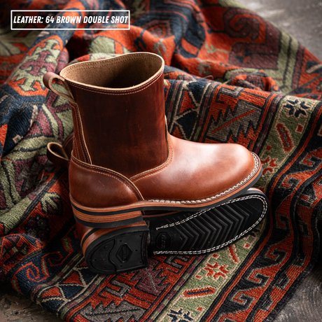 A pair of brown leather ankle boots displayed on a patterned red, green, and black rug. One boot is standing upright, showing its smooth brown leather shaft and pull tab, while the other lies on its side, displaying the black treaded sole with white stitching.