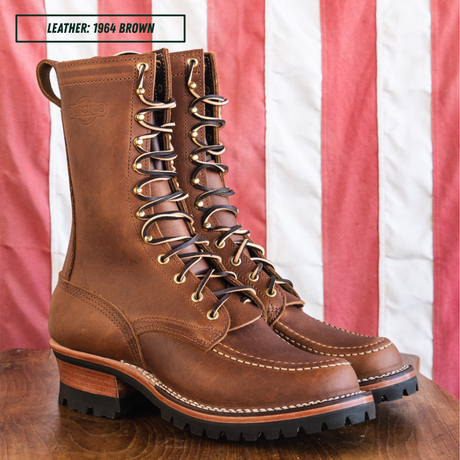 A pair of brown leather lace-up boots displayed on a wooden surface with a red and white striped fabric backdrop. A text label in the top left reads 'LEATHER: 1964 BROWN'.