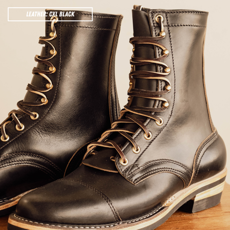 A pair of black leather lace-up ankle boots with gold-toned metal eyelets and laces, shown on a wooden surface against a light beige background. A text overlay in the top left reads 'LEATHER: CXL BLACK'.